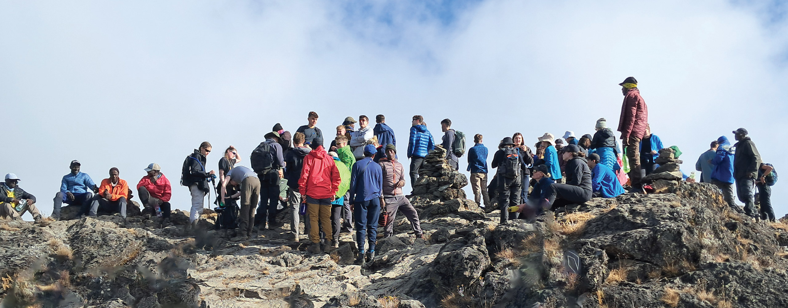 A large group of hikers in outdoor gear gathered on a rocky mountain summit under a partly cloudy sky
