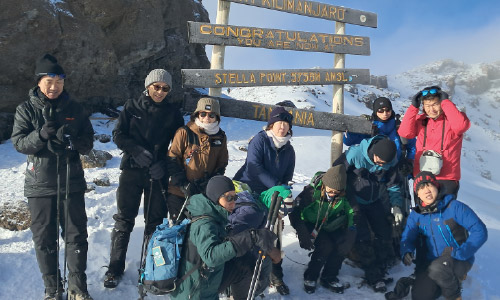 Eleven climbers in winter gear pose in front of the Stella Point sign on snowy Mount Kilimanjaro.
